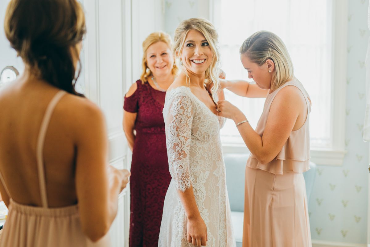 Bridesmaids helping smiling bride adjust her lace wedding dress before the ceremony.