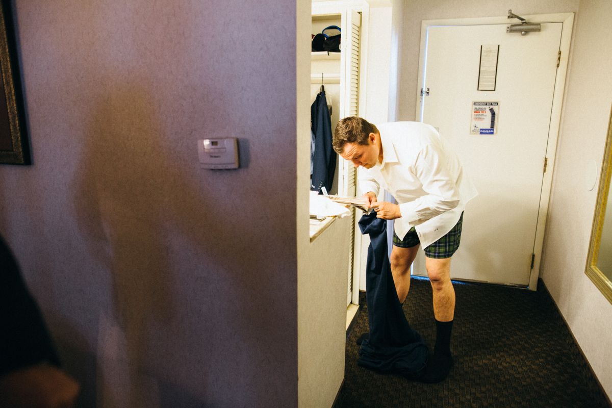 Groomsman in kilt adjusting outfit in hotel hallway while preparing for wedding.