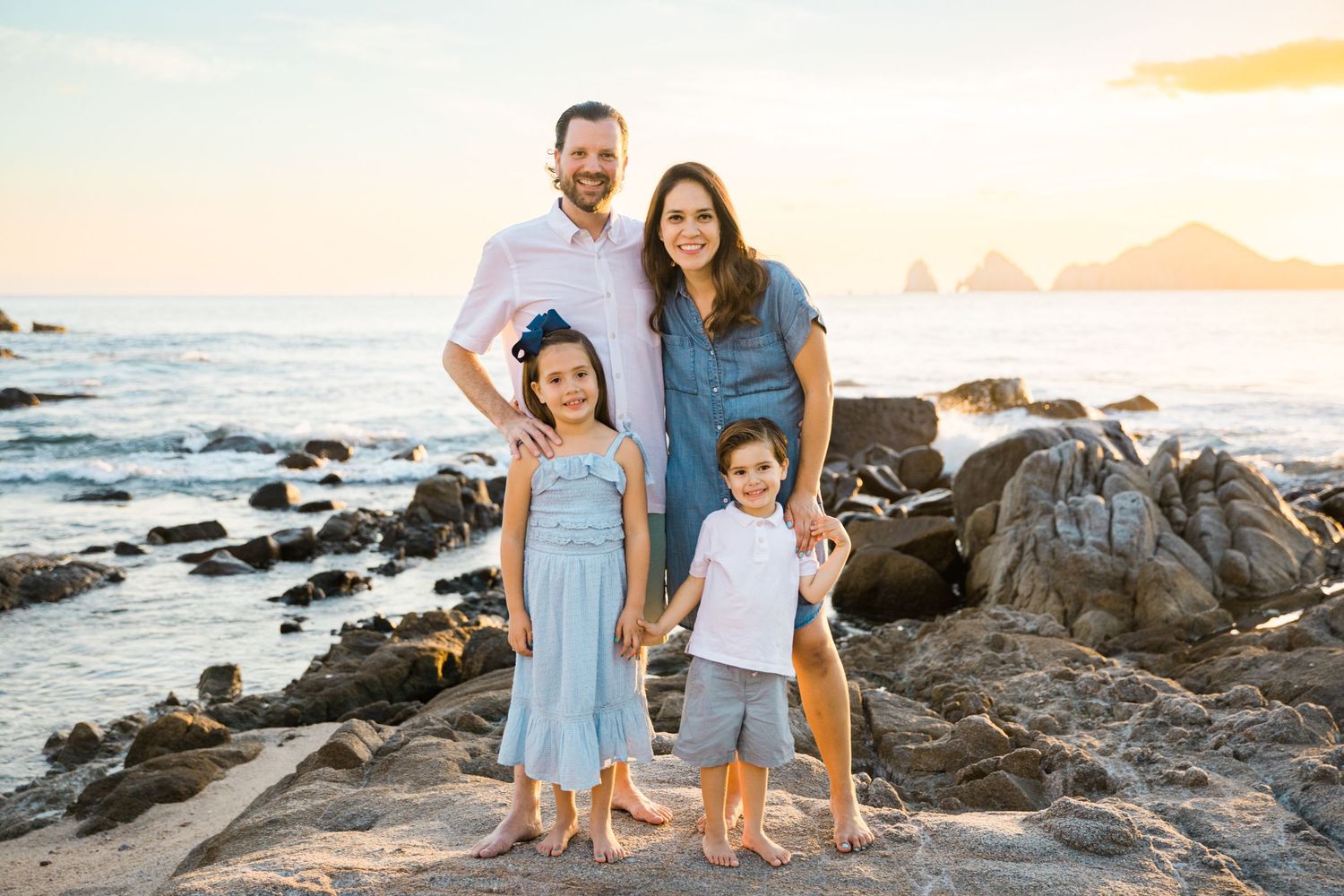 Family of four smiling on rocky beach at sunset with ocean and mountains in background in Cabo San Lucas