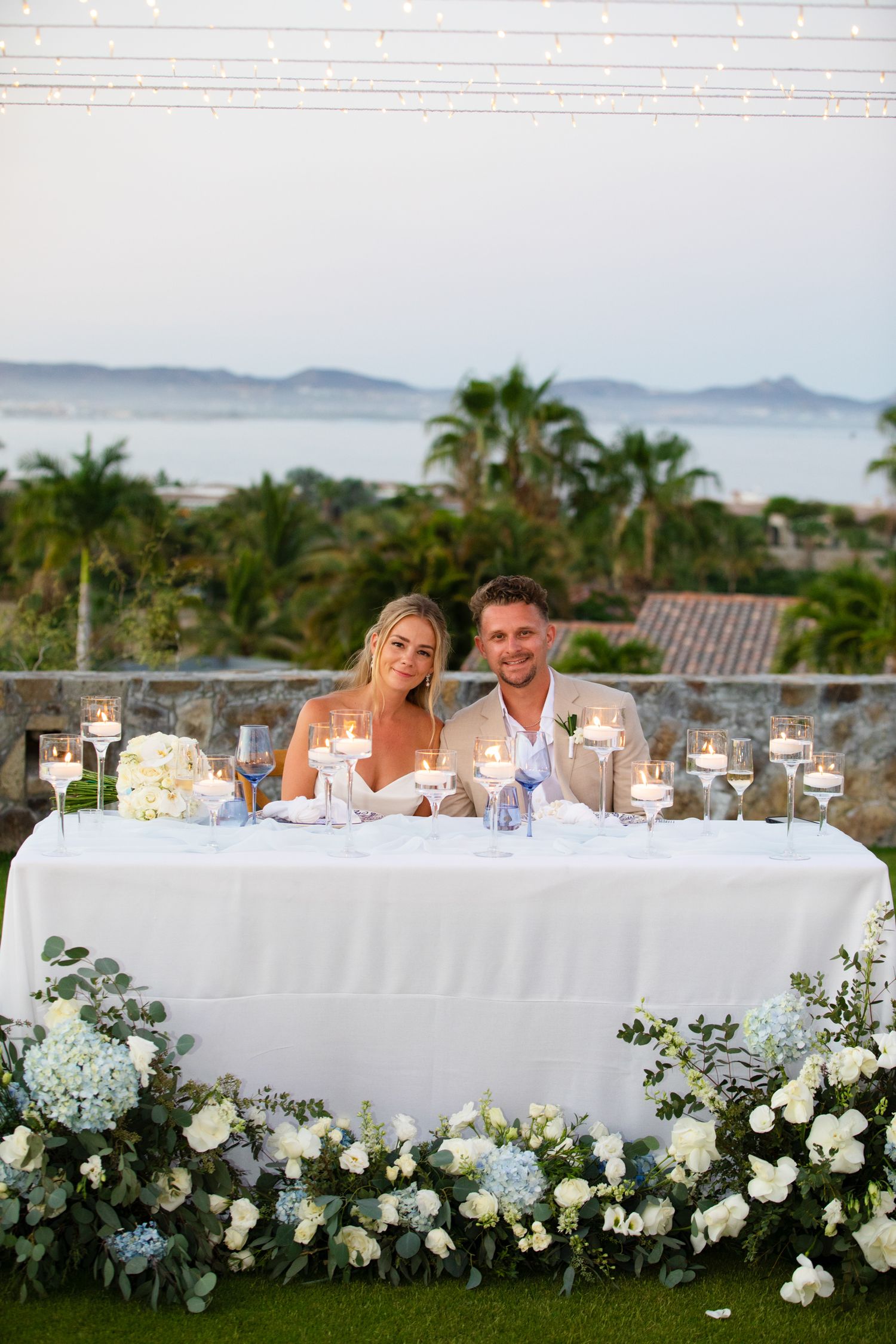 Bride and groom seated at floral-decorated sweetheart table with candles and ocean view backdrop at outdoor wedding reception.