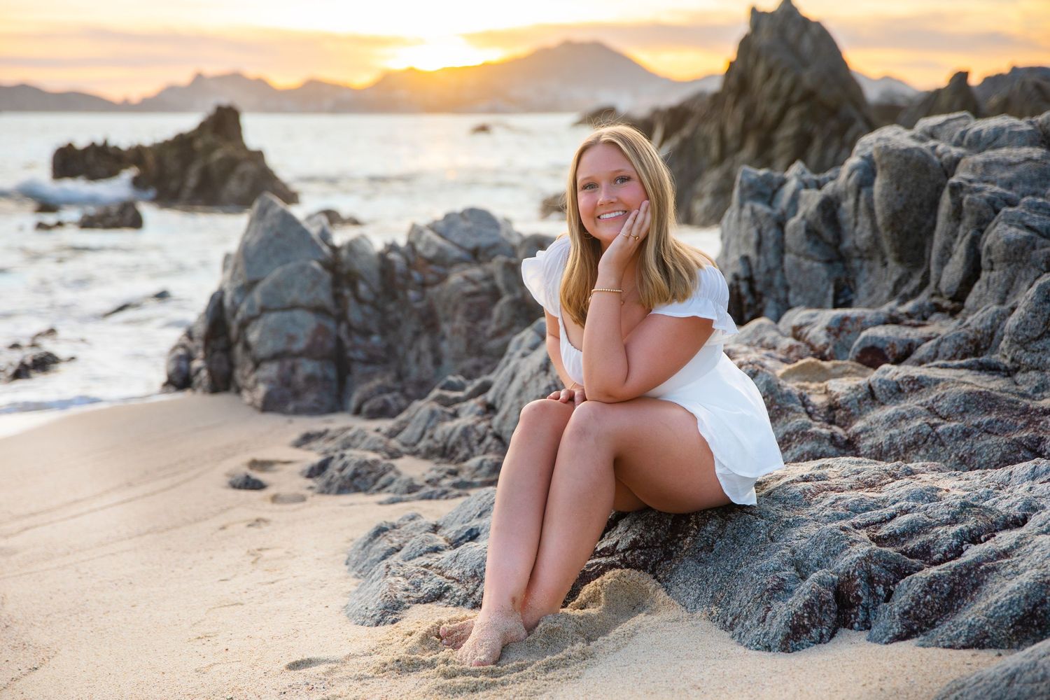 Young woman in white dress sitting on rocks at a scenic beach during golden hour sunset.