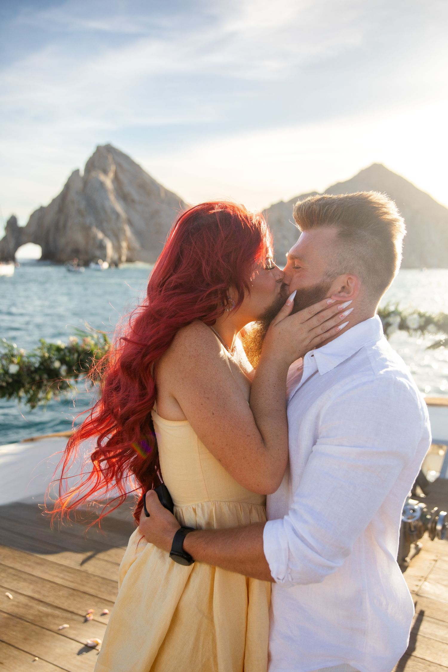 Couple kissing on a boat deck with iconic Cabo San Lucas arch and ocean in the background at sunset.