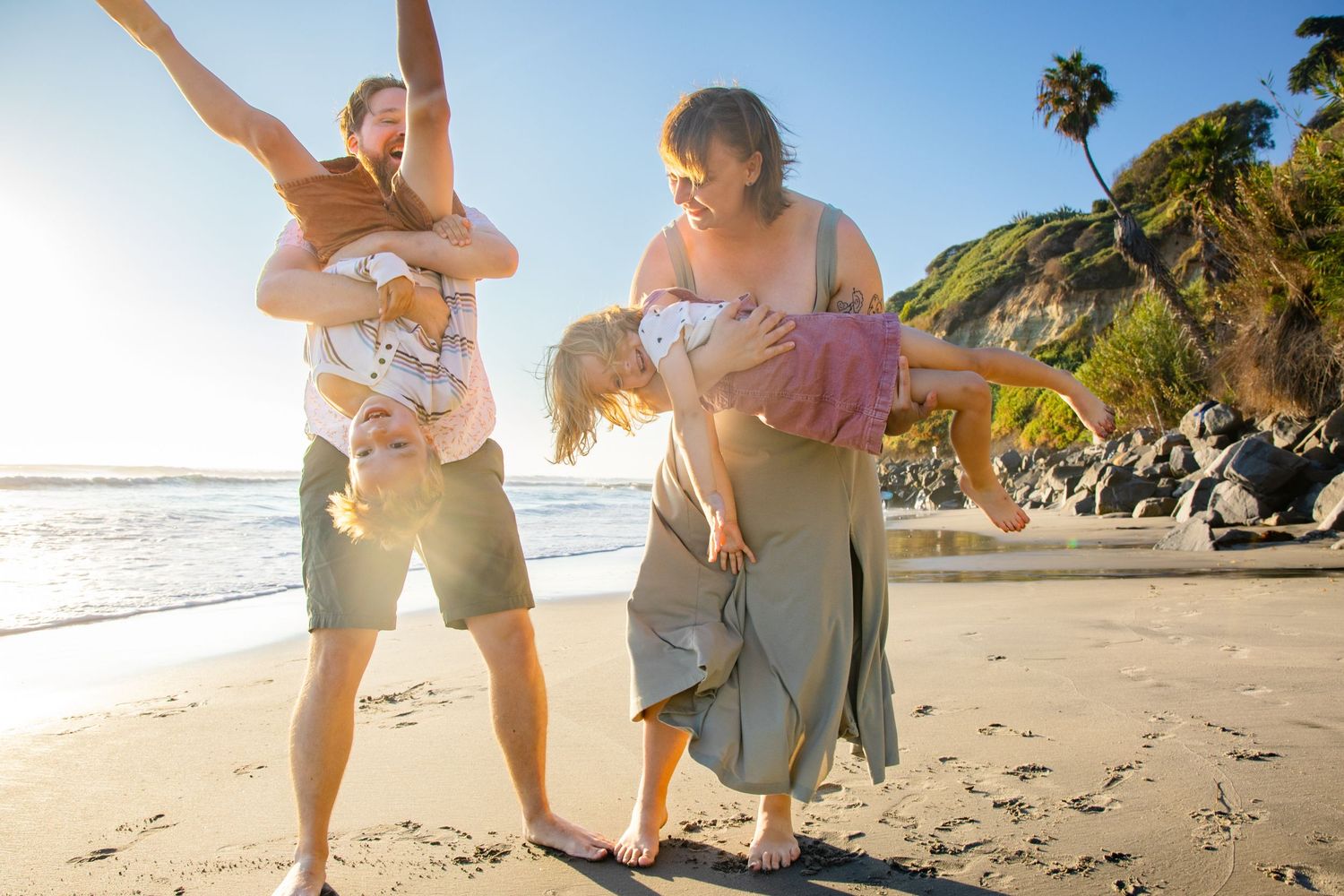 Family of four playing on a sunny beach, parents playfully lifting children with ocean waves and palm trees in background in Cabo San Lucas