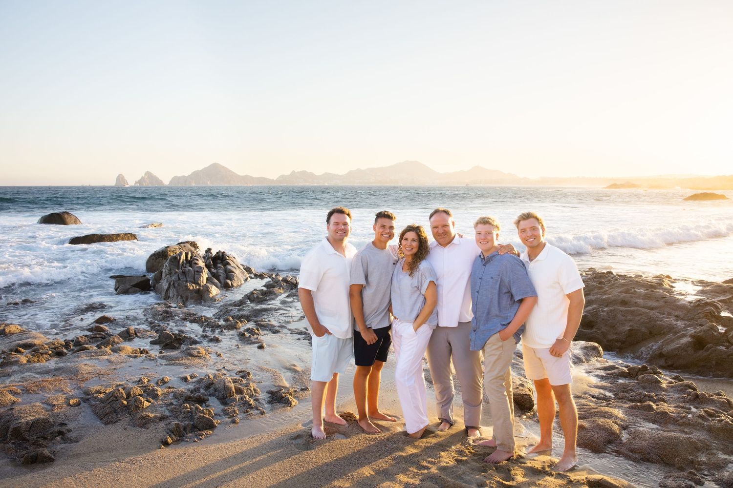 Large group of seven adults posing together on rocky beach during golden sunset.