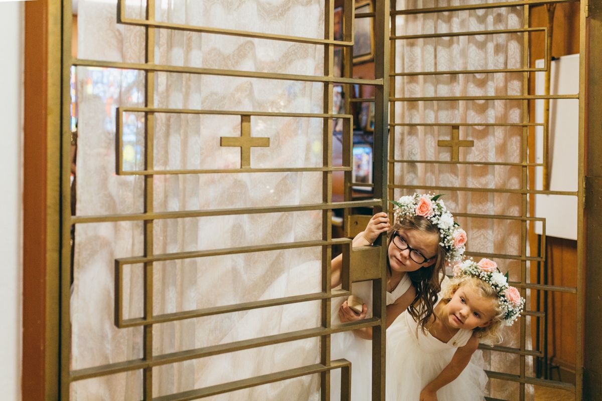 Bride and flower girl peeking playfully through decorative gold gate at church wedding.