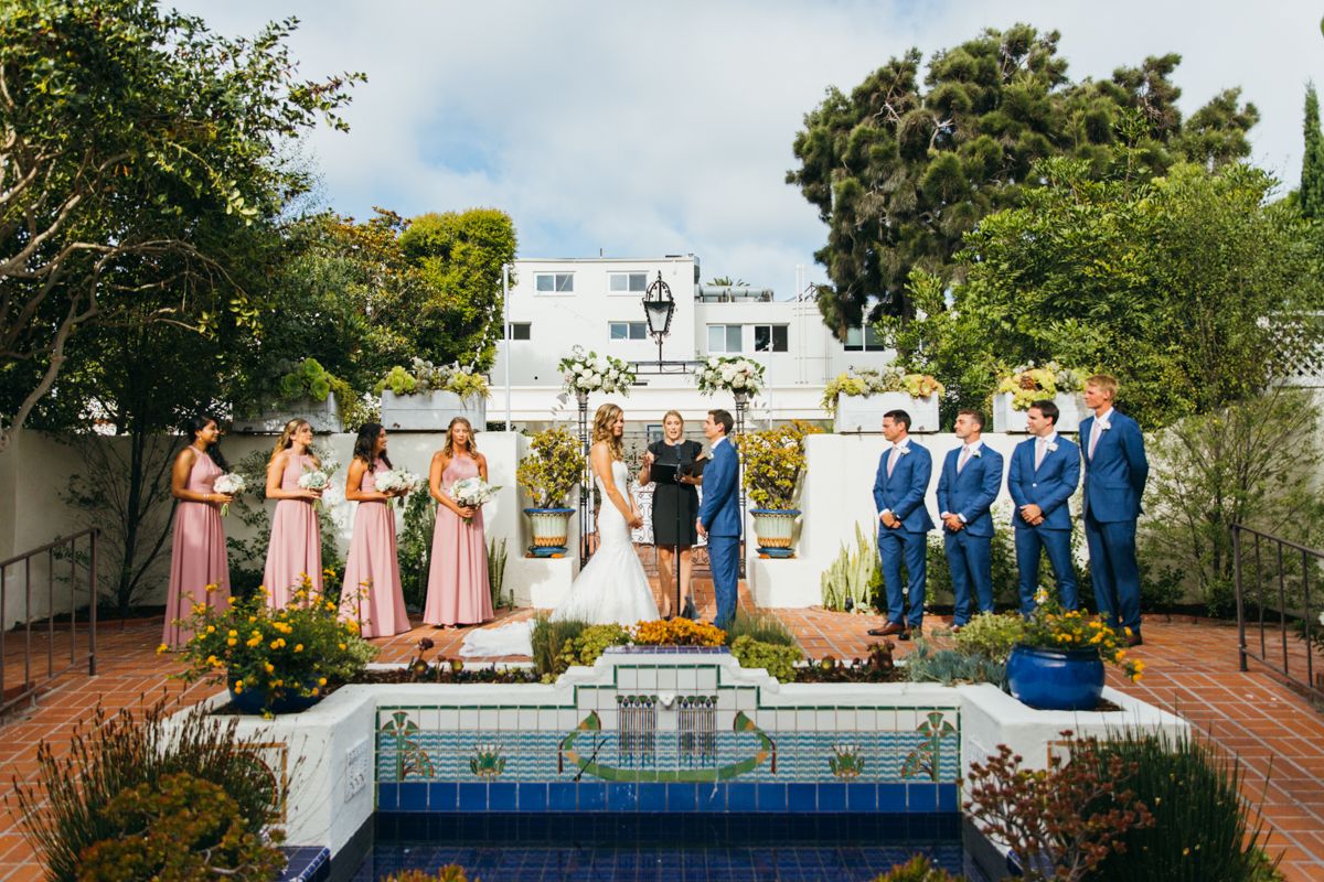 Wedding party stands near a decorative tiled fountain in a lush garden during an outdoor ceremony.