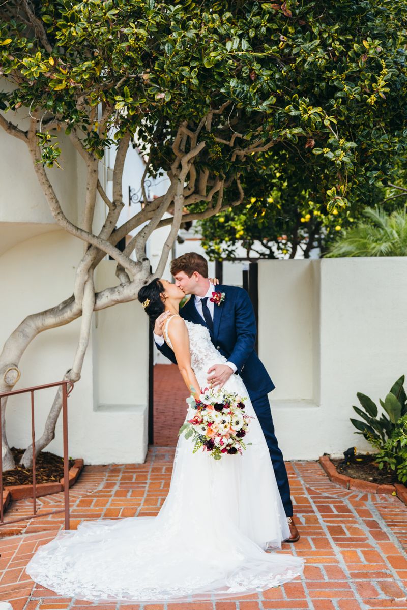 Bride and groom share a romantic kiss under a lush green tree on a brick patio on their wedding day.