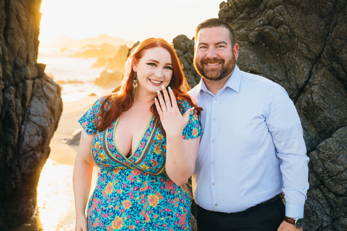 Happy couple showing engagement ring on beach at sunset, woman in floral dress smiling at camera.