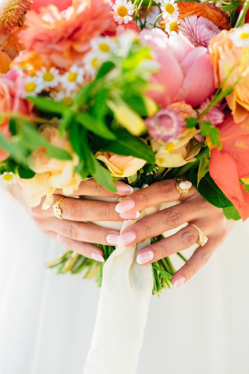 Bride holds vibrant bouquet of pink, orange, and white flowers with gold wedding rings.