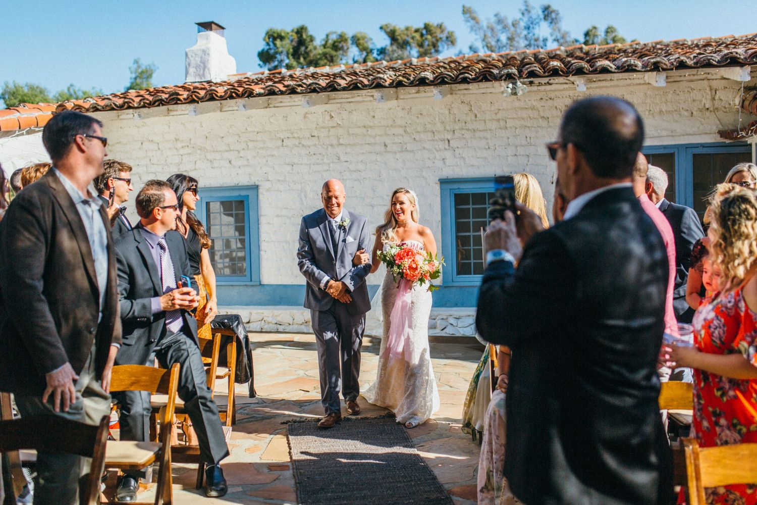 Bride walks down outdoor aisle with escort at sunny wedding ceremony,  beside Spanish-style building Leo Carrillo ranch wedding