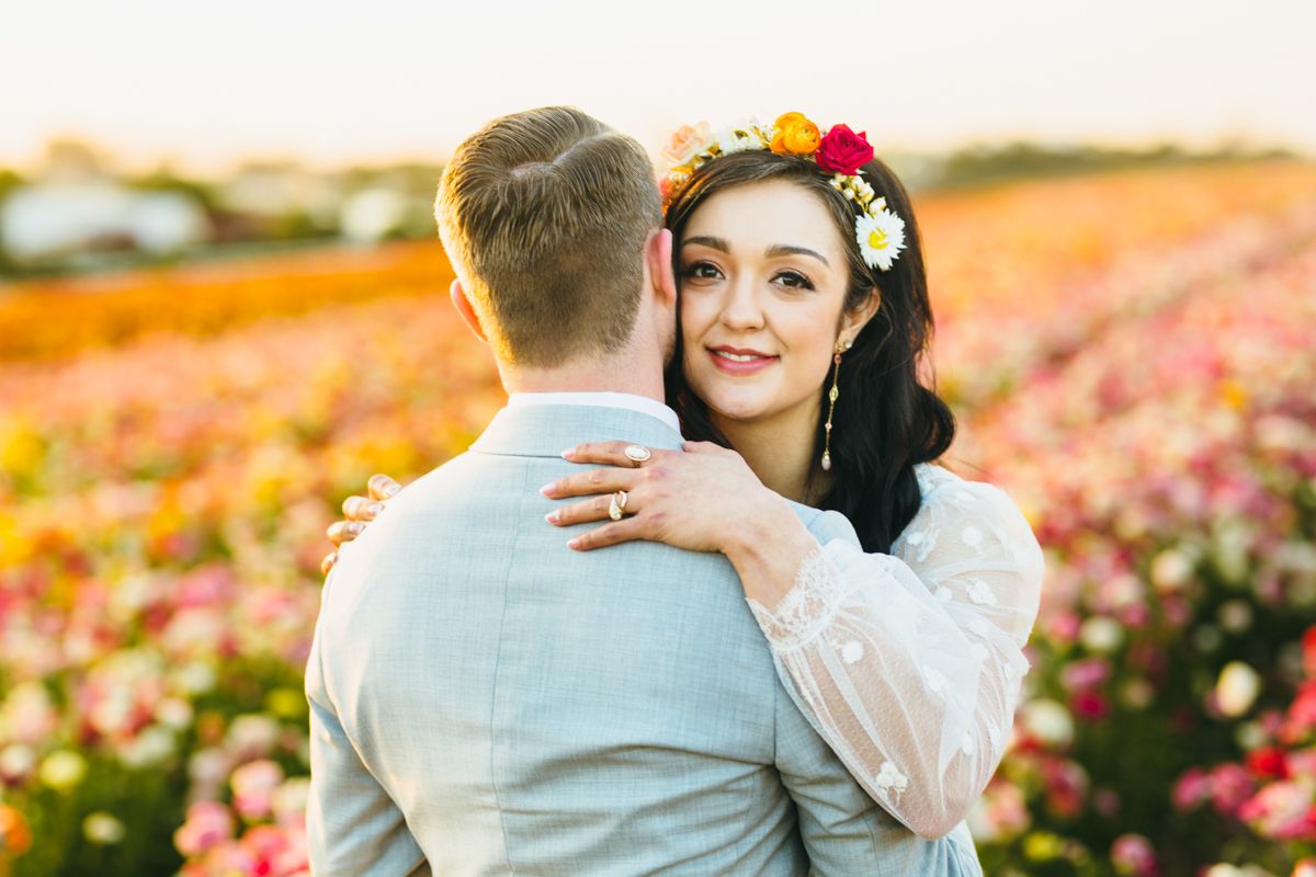 Couple embraces in colorful flower field, bride wearing floral crown smiles over groom's shoulder at sunset.