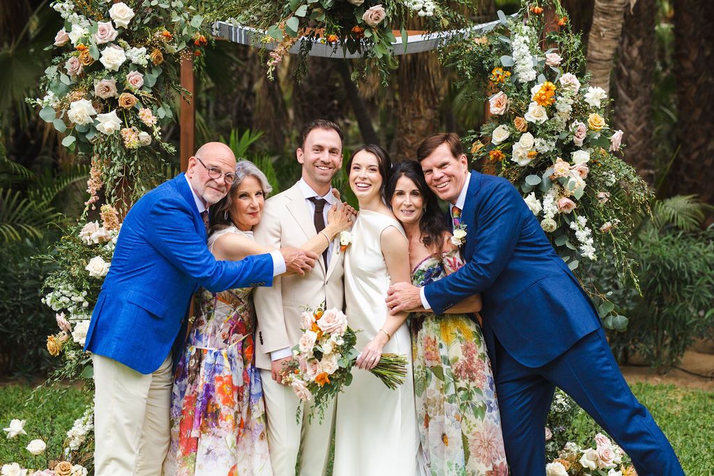 A joyful wedding party poses together in front of a stunning floral arch at an outdoor garden wedding reception.