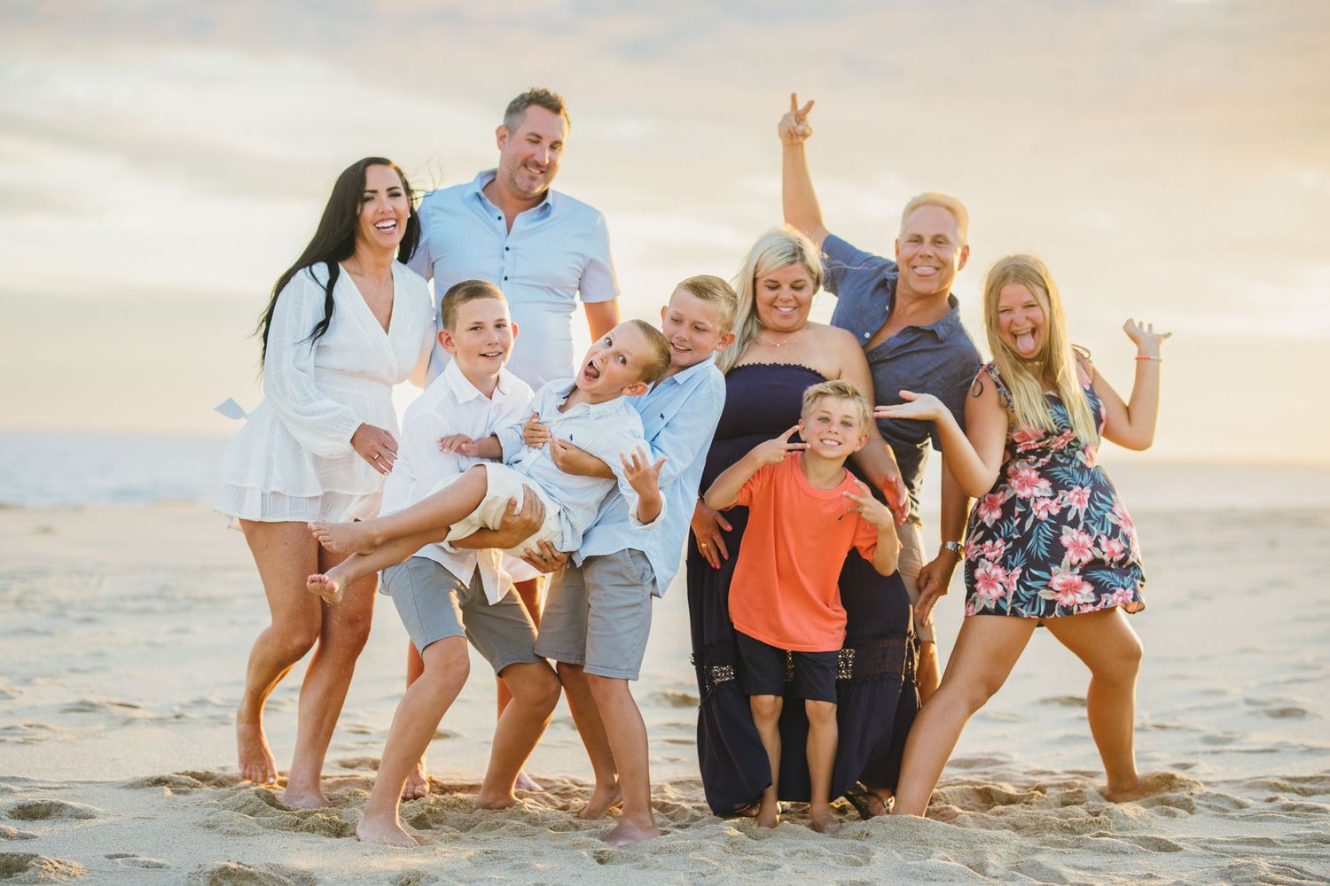 A large family poses playfully on a sandy beach in Cabo San Lucas at sunset, wearing coordinated light-colored outfits.
