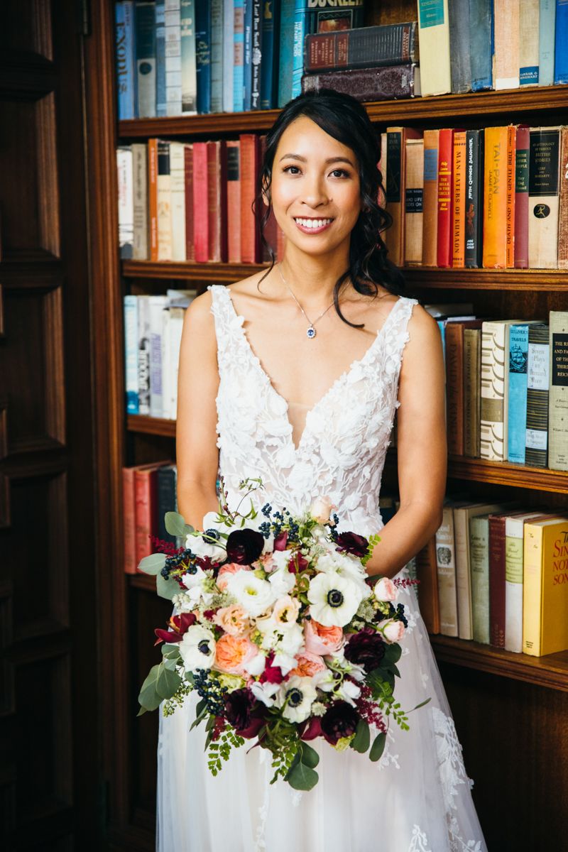 Smiling bride in lace wedding dress holding a colorful bouquet in front of a bookshelf.