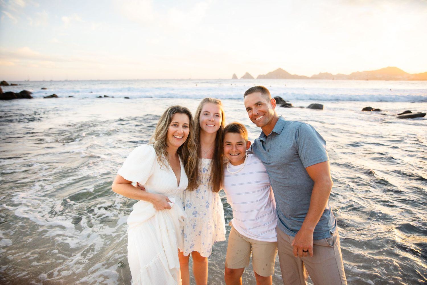 Family of four smiling together on a sandy beach at sunset  in Cabo San Lucas with ocean waves and rocky coastline in background.