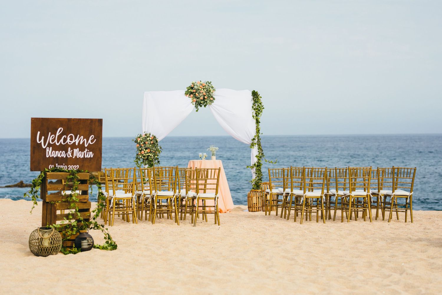 Beach wedding setup in cabo with gold chairs, white floral arch, and welcome sign by the ocean.
