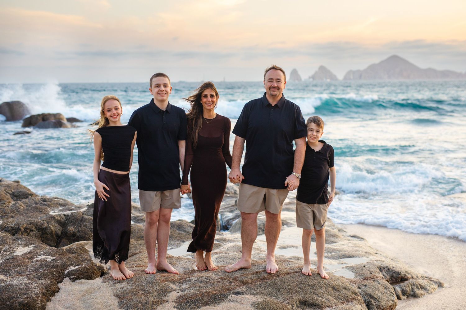 Family of five posing on rocky beach at sunset with ocean waves and scenic coastline in background in Cabo San Lucas