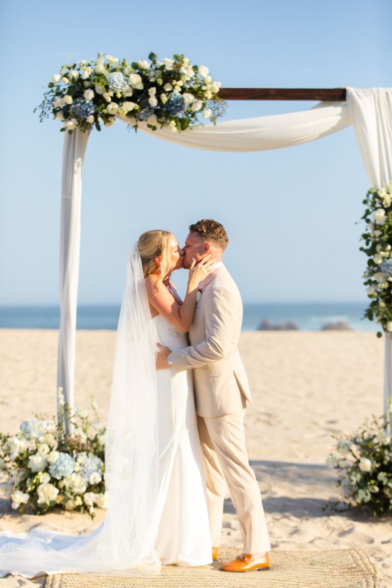 Bride and groom share first kiss under floral arch adorned with blue and white flowers.