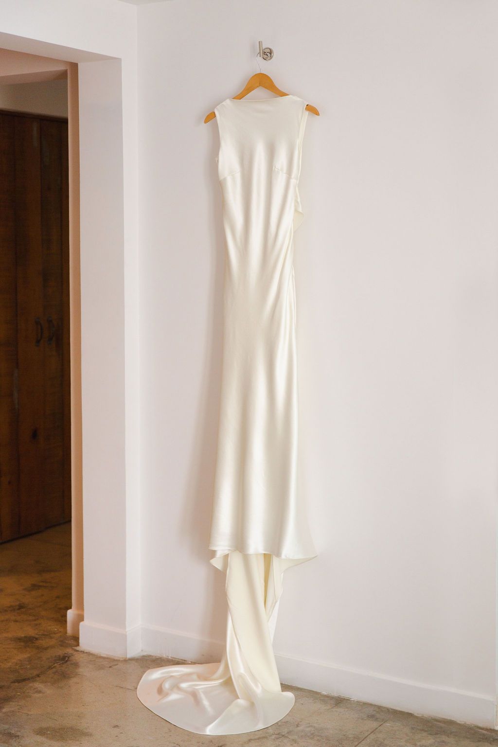 Minimalist sleek white satin wedding gown with a train hanging on a wooden hanger against a white wall.