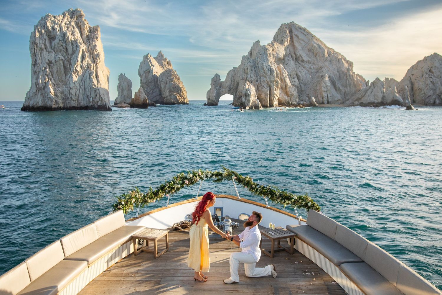 Man proposes to woman on decorated yacht bow with iconic Arch of Cabo San Lucas rock formations in background.