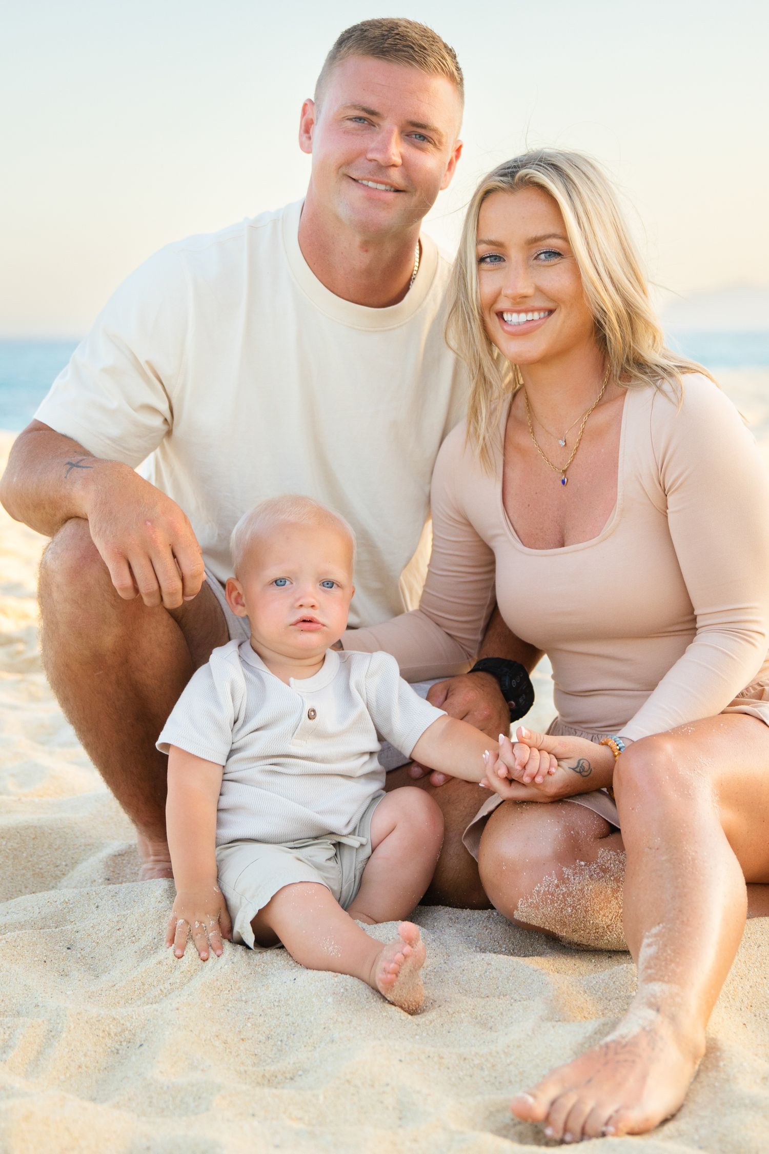 Happy family of three posing on sandy beach at sunset, parents smiling with baby boy sitting between them.