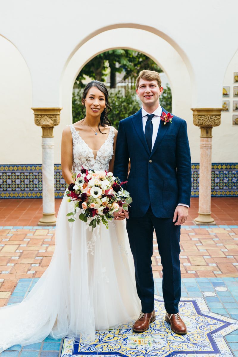 Bride and groom posing together under arched columns holding a colorful floral wedding bouquet.