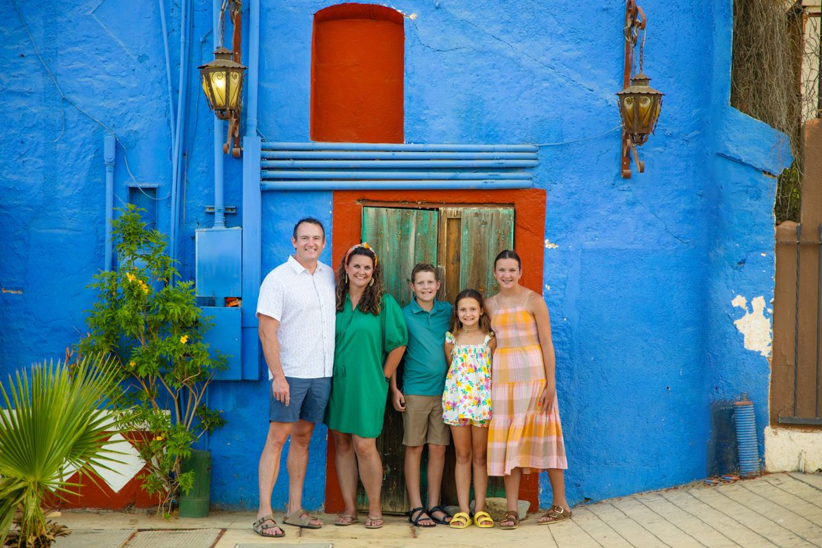 Family of five smiling together in front of bright blue wall with red door.