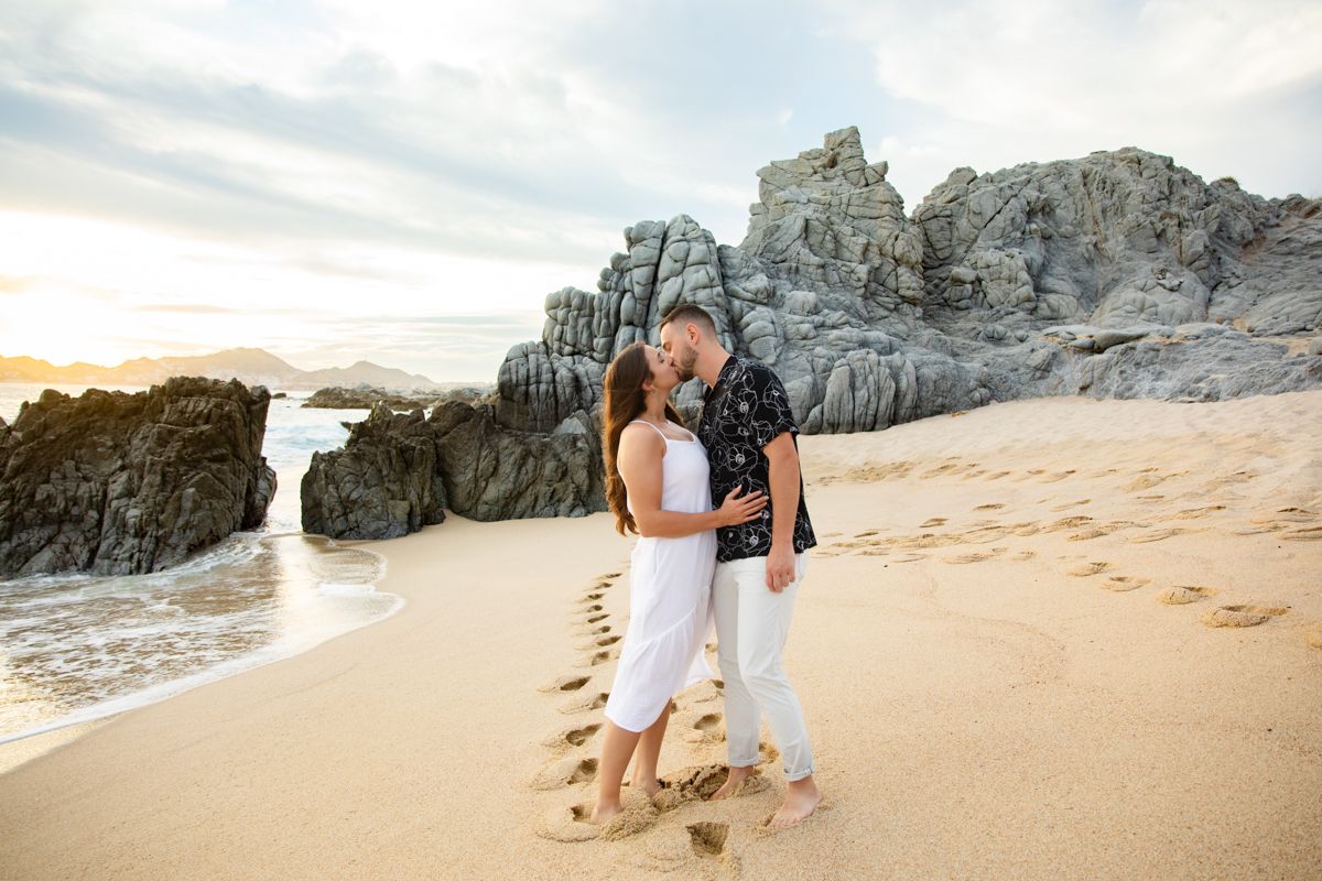 Couple embraces and kisses on sandy beach at golden sunset with dramatic rock formations nearby.