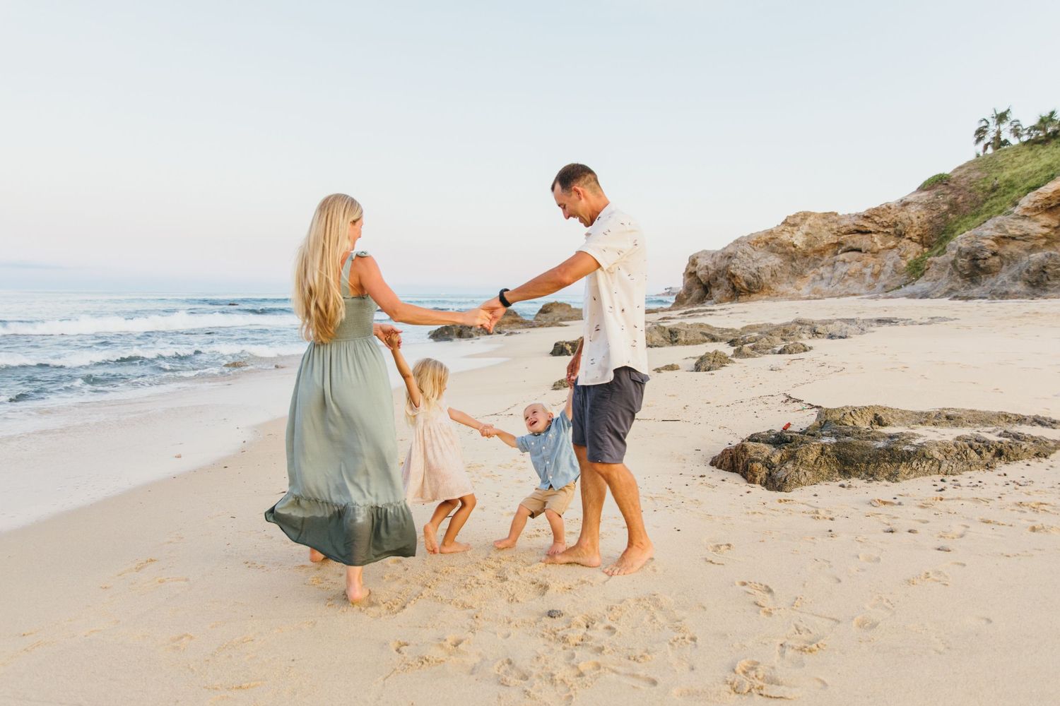 Family of four holding hands and dancing on a sandy beach at sunset with ocean waves in the background.