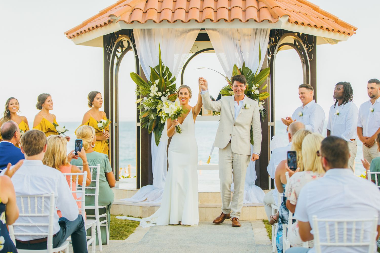 Bride and groom celebrate at outdoor beach wedding ceremony under elegant gazebo with tropical floral arrangements.