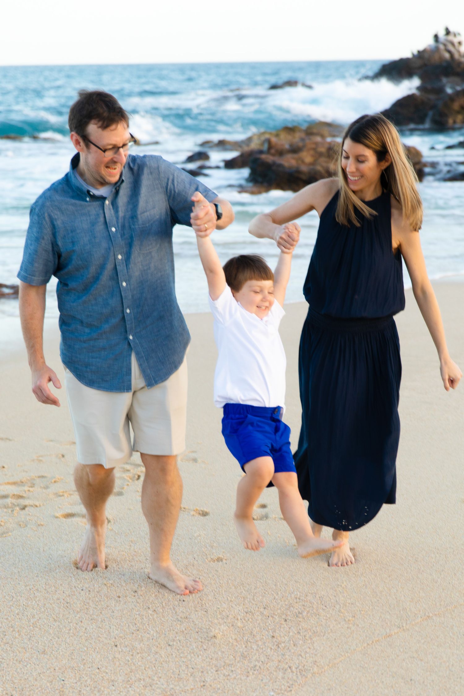 Family of three enjoying a sunny beach day, parents swinging young boy by his hands on sandy shore in Cabo San Lucas.