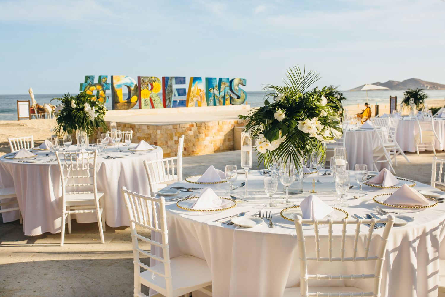 Elegant outdoor beach wedding reception with white tables, floral centerpieces, and colorful Dreams resort sign backdrop.
