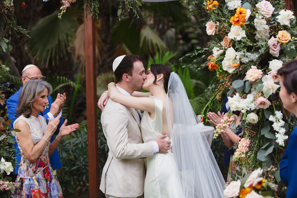 Bride and groom sharing their first kiss at an outdoor wedding ceremony with floral arch.