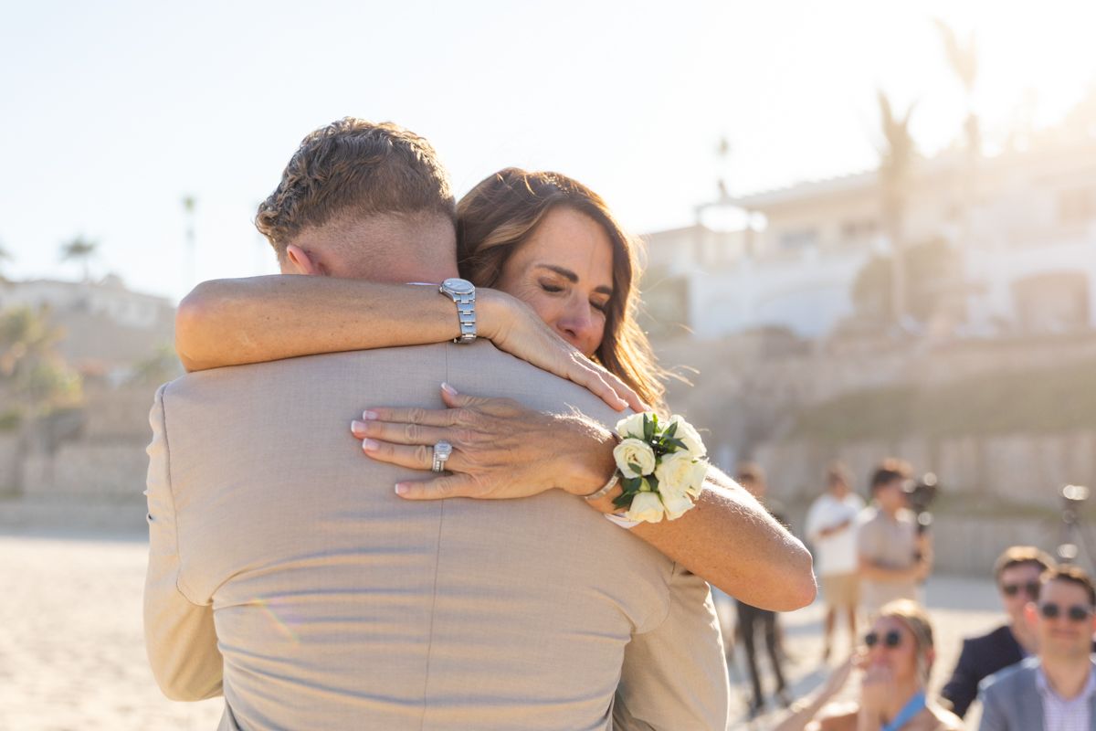 Emotional bride hugs groom on beach during wedding ceremony with guests in background.