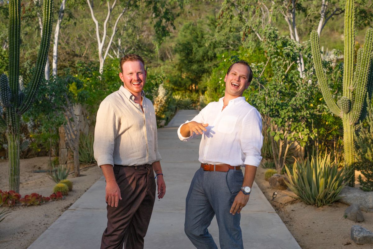 Two men laughing and showing engagement ring on a sunlit desert garden pathway with saguaro cacti.