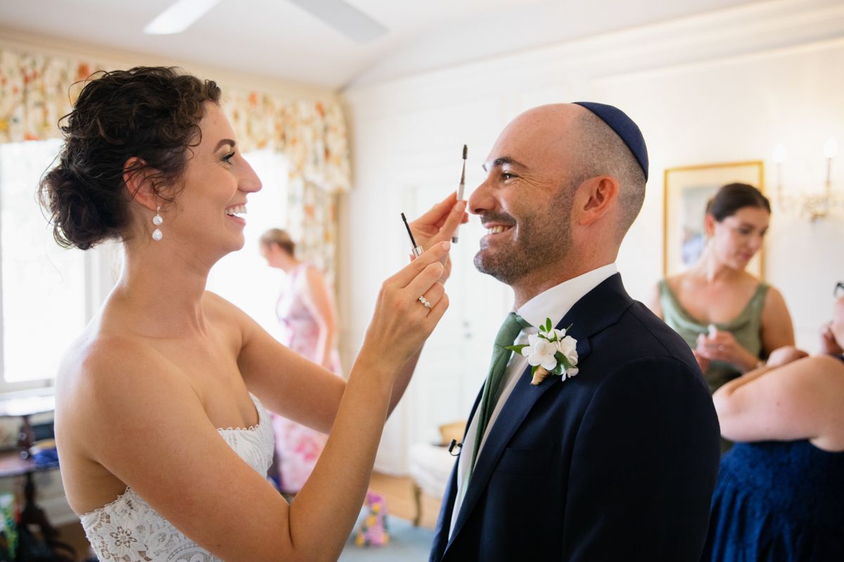 Bride applies makeup to smiling groom wearing kippah in navy suit with floral boutonniere before Jewish wedding.