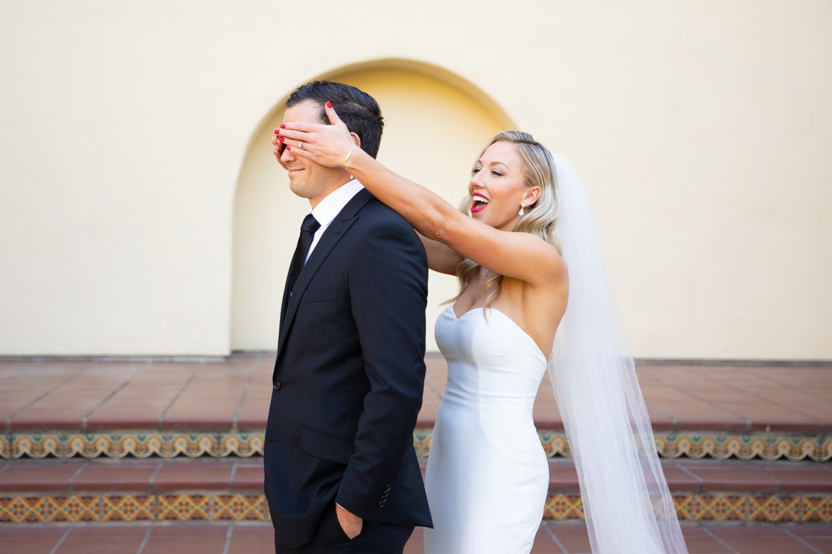 Bride surprises groom by covering his eyes during fun wedding first look moment outdoors.