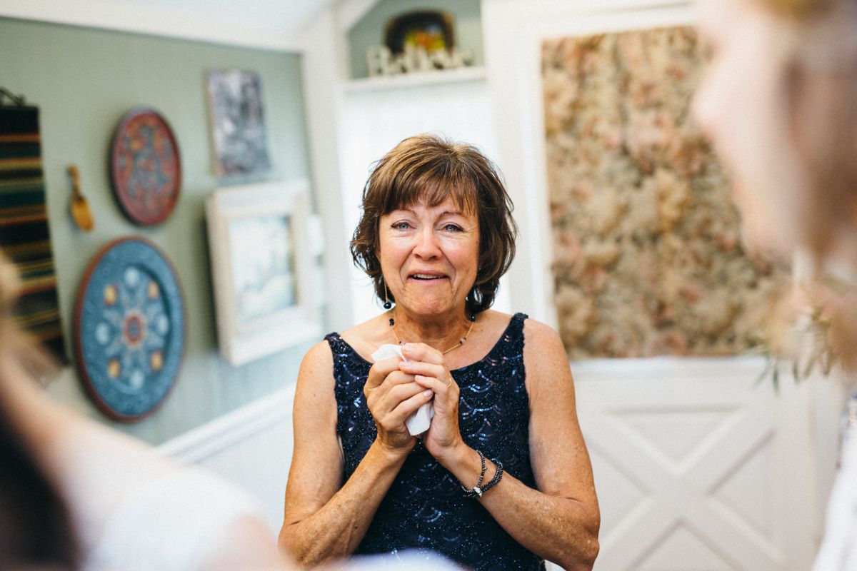 An elegant older woman clasps her hands and smiles warmly during a wedding celebration indoors.