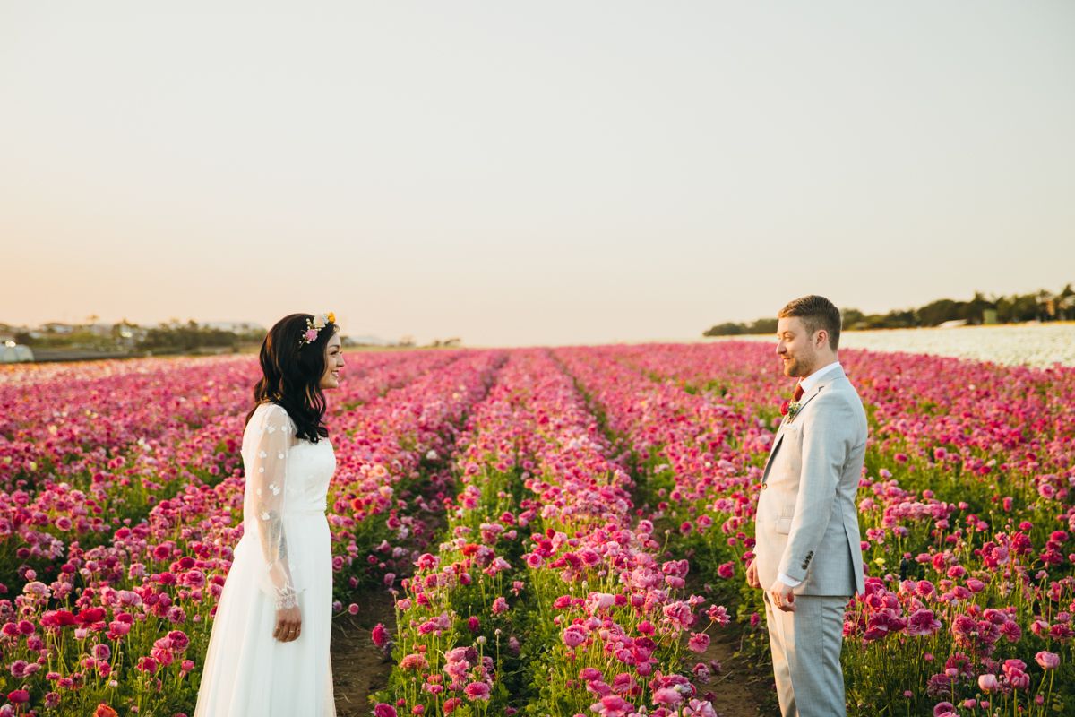 A bride and groom face each other in a stunning pink flower field at sunset.