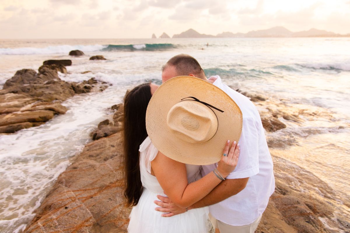 Couple sharing a romantic kiss on rocky shoreline at sunset, woman wearing a wide-brim straw hat.