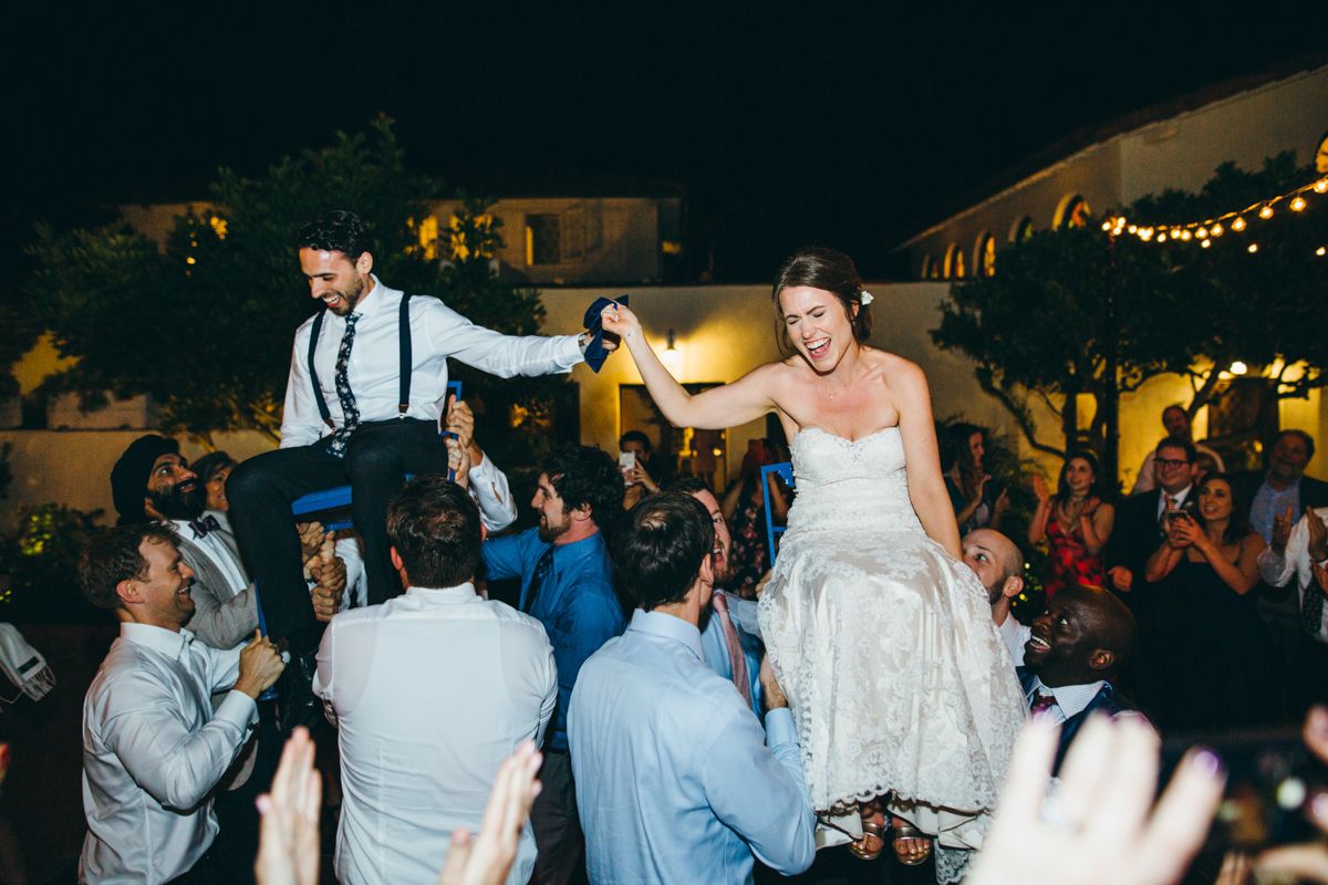 Bride and groom lifted by guests during energetic hora dance at nighttime wedding reception.