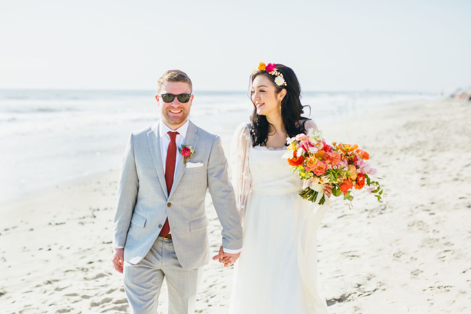 Bride and groom walking hand in hand along a sunny beach on their wedding day.