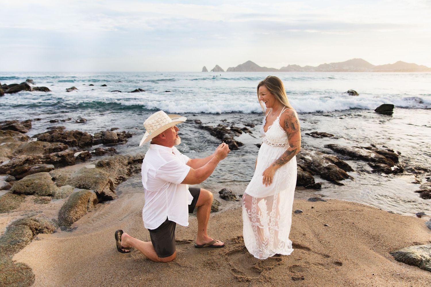 Man proposing to woman on a rocky beach at sunset, holding a ring while kneeling in sand near ocean waves.