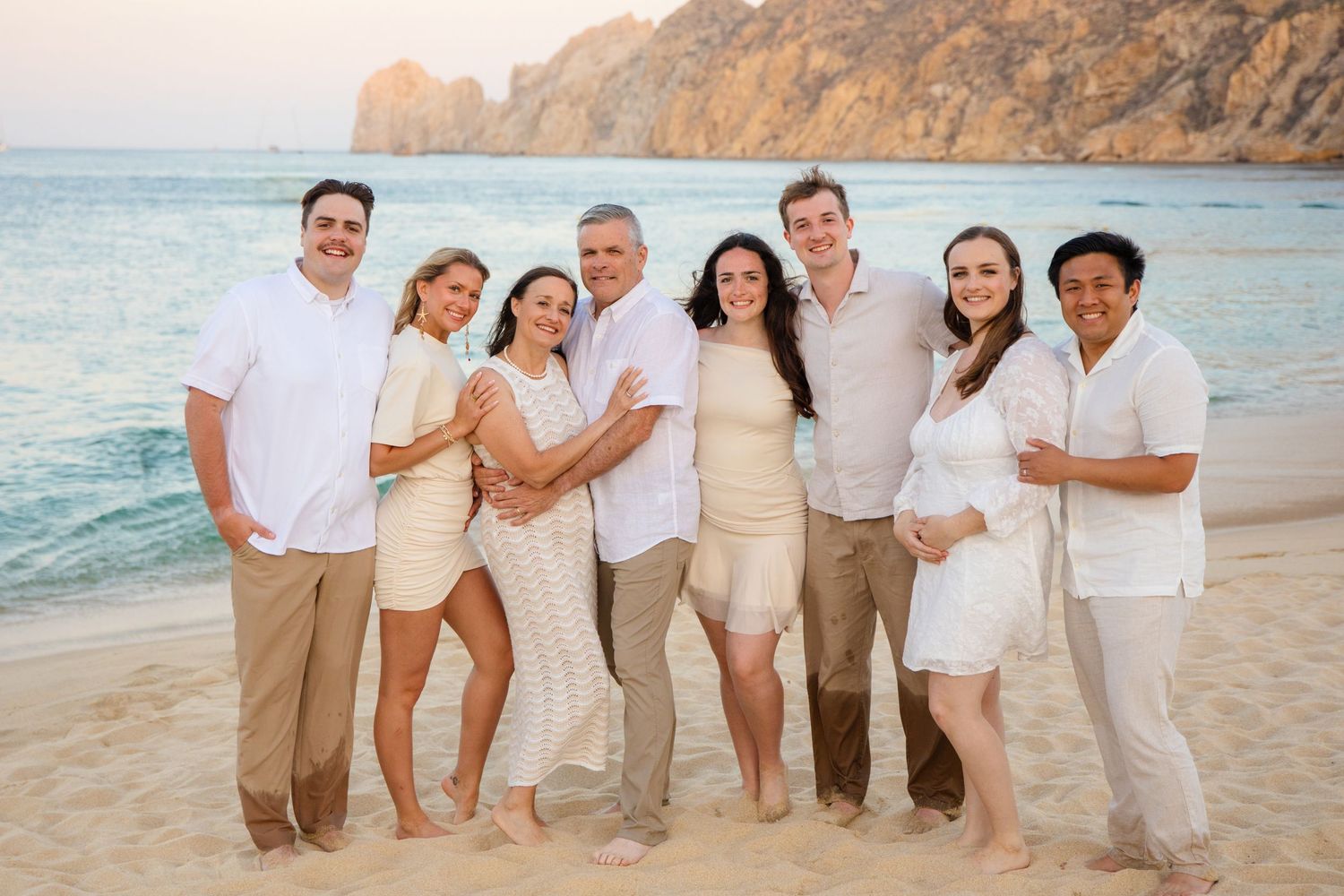 Happy family of seven posing together on a sandy beach at sunset with rocky cliffs in the background in Cabo San Lucas Mexico.