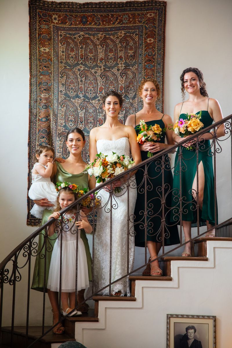 Bride with bridesmaids in green dresses and flower girls pose on ornate staircase before tapestry.