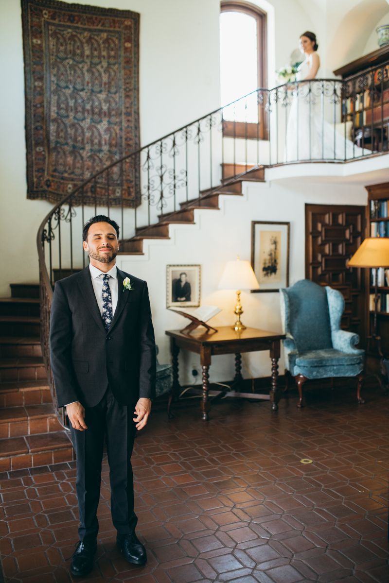 Groom in dark suit standing by ornate staircase with bride visible on balcony above in historic venue.