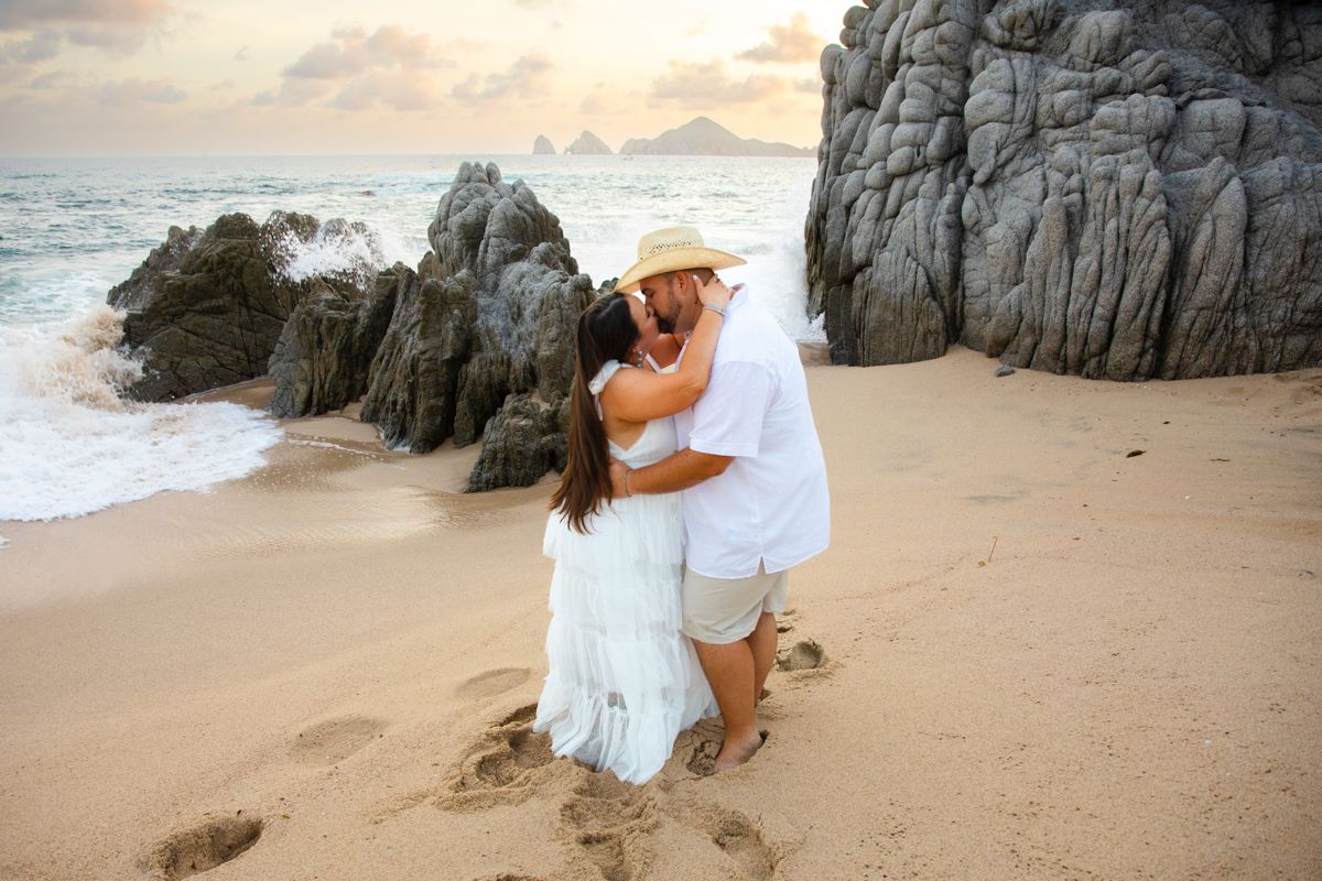 Couple embracing and kissing on sandy beach at golden hour with dramatic rock formations in background.