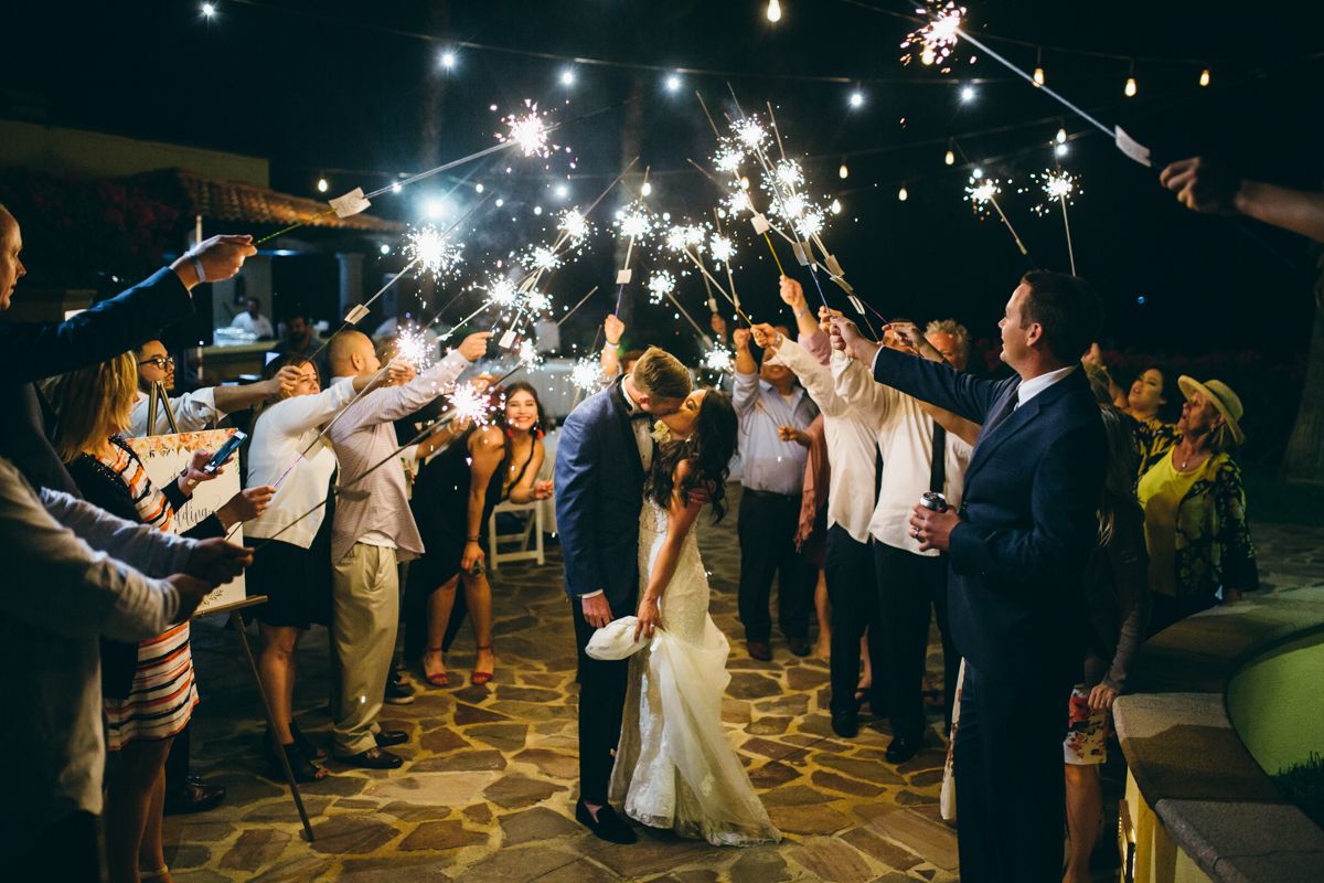Newlyweds share kiss surrounded by guests holding sparklers during magical nighttime wedding sendoff.