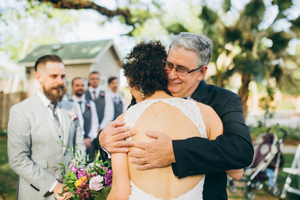 Emotional bride hugs older man while groomsmen watch during outdoor wedding ceremony.