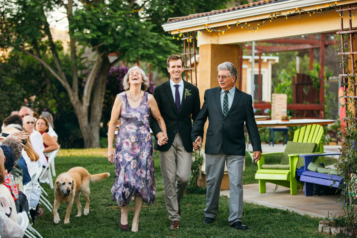 Groom walks down outdoor aisle with smiling parents and golden retriever dog at backyard wedding ceremony.
